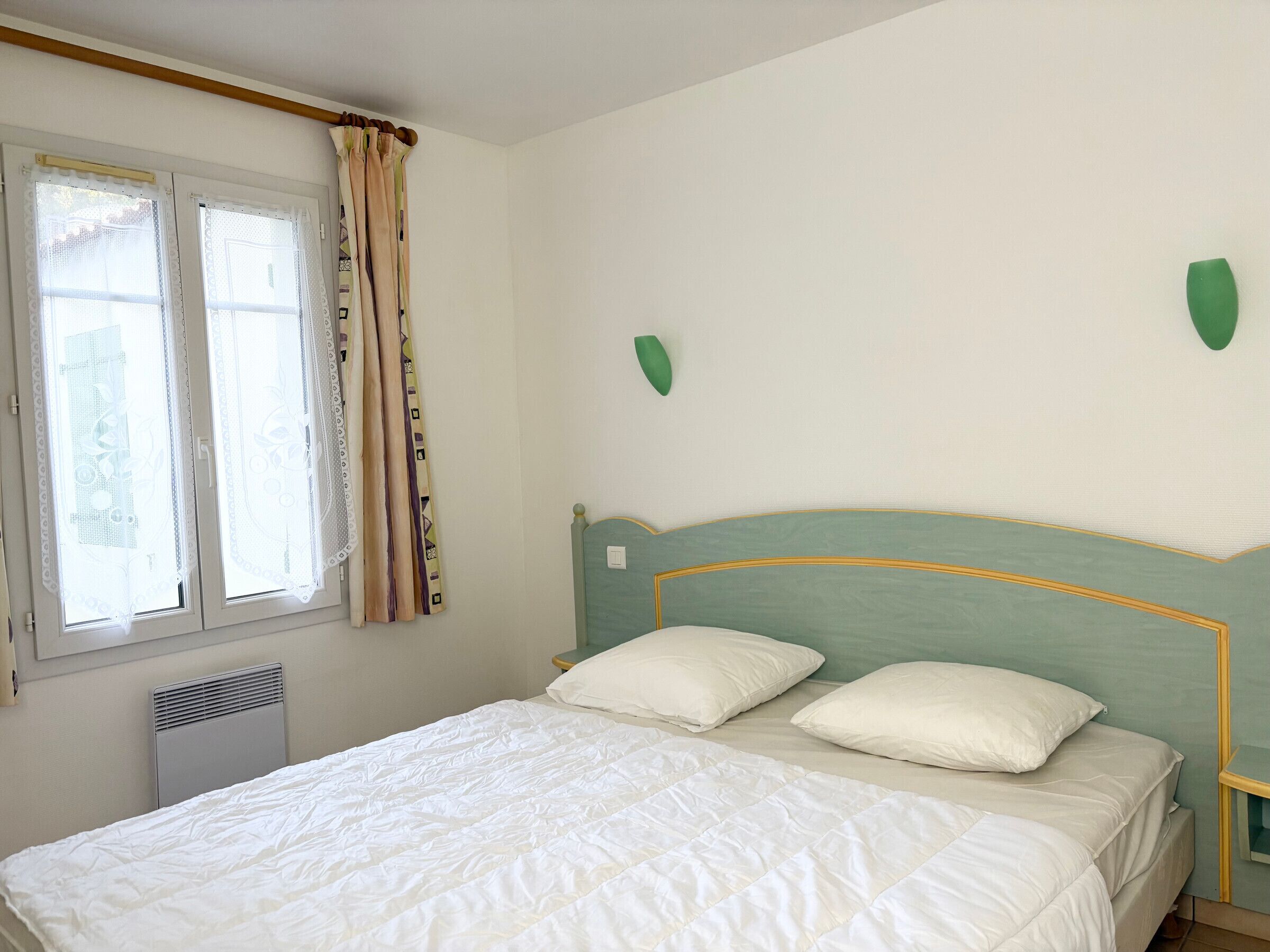 Ein Schlafzimmer mit einem Bett, zwei Kissen, einem Fenster mit Vorhängen, einer Heizung und einer Wanddekoration in Grün.