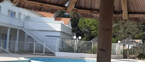 A swimming pool with a tiled edge, a wooden pergola overhead, and a white building with a staircase.