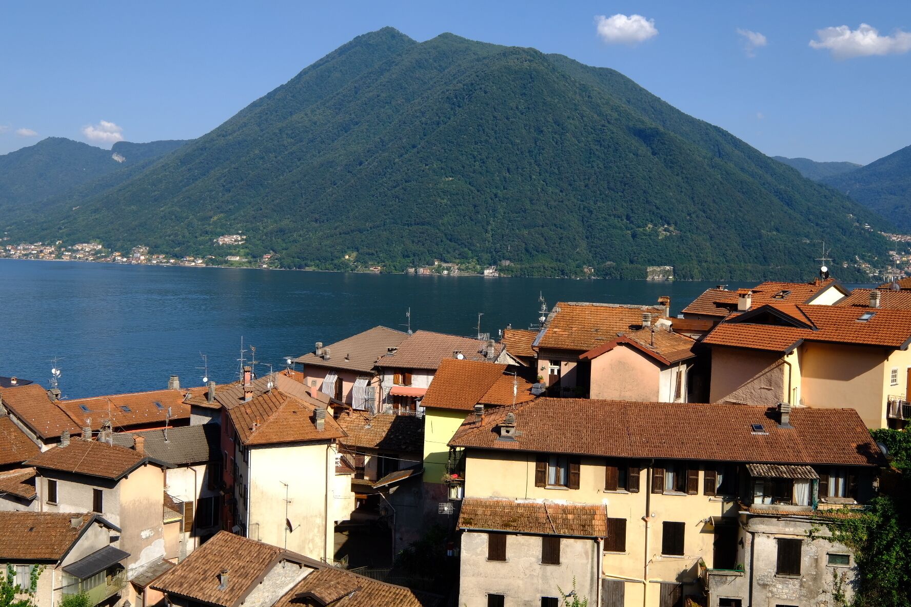 A coastal town with terracotta-roofed buildings, a mountain in the background, and a body of water in the foreground.