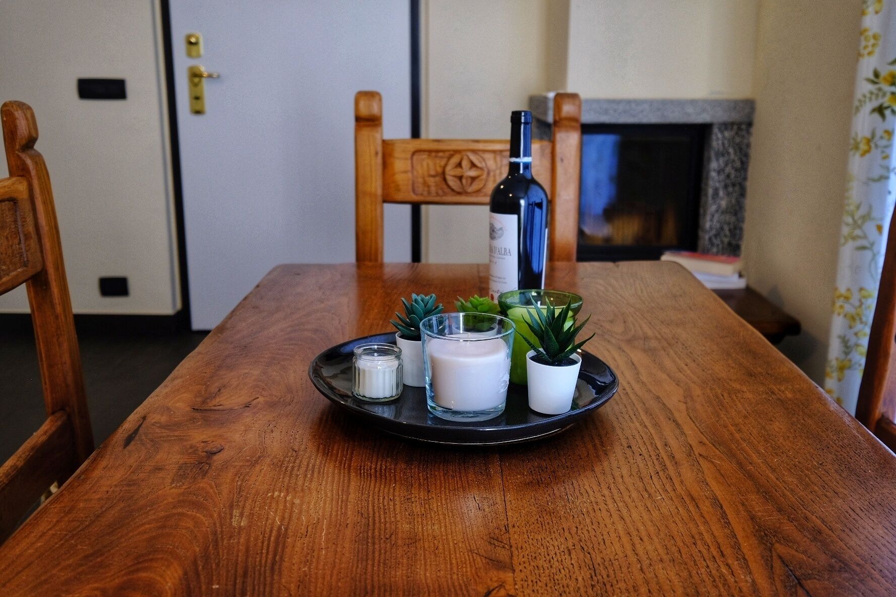 A wooden dining table with a black tray holding a bottle, candles, and small potted plants.
