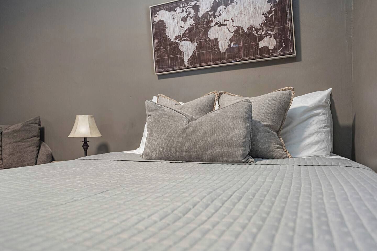 A neatly made bed with a quilted grey bedspread and grey pillows, a world map on the wall, and a bedside lamp.