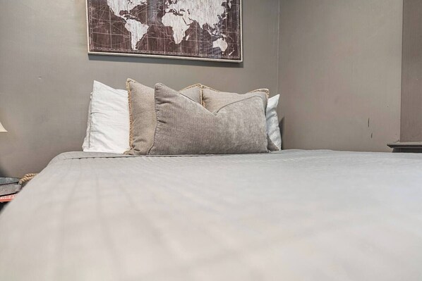 Una cama bien hecha con un edredón gris y dos almohadas, un mapa mundial en la pared y una mesita con una lámpara.