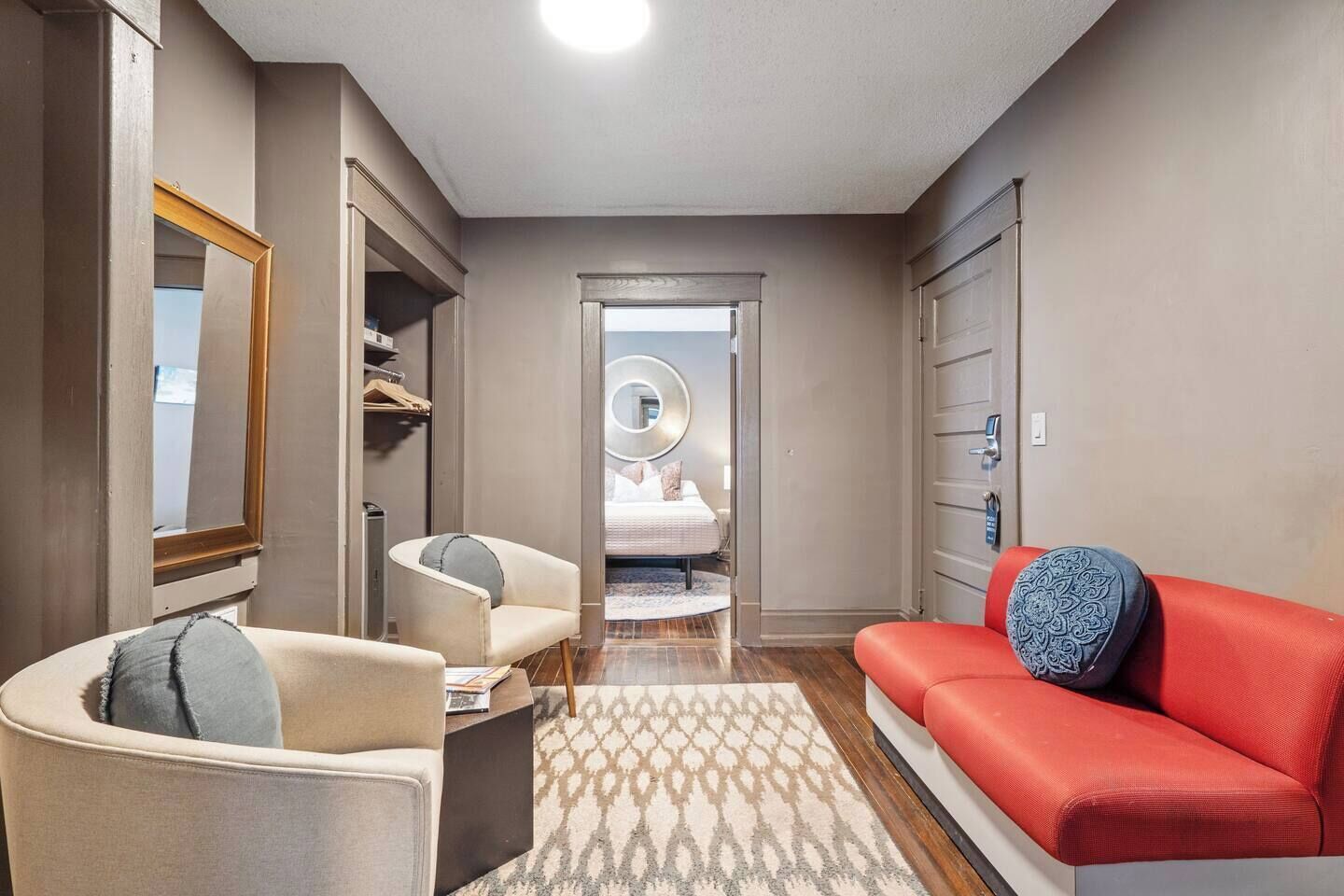 A modern hotel room with a bed, a red sofa, a chair, a mirror, and a wardrobe.