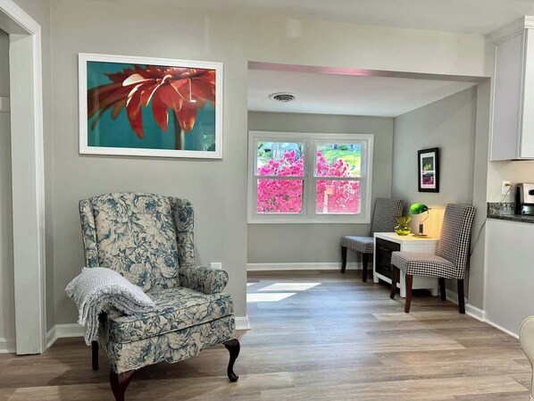 Un salon avec un fauteuil à motifs floraux, un coin repas avec des chaises à carreaux et une fenêtre ornée de fleurs roses.