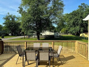 A wooden deck with a table and chairs, surrounded by a wooden railing, overlooking a grassy area with trees and a distant road. - Family Home on Mayo Peninsula | Deck | Sleeps 10 (Edgewater)
