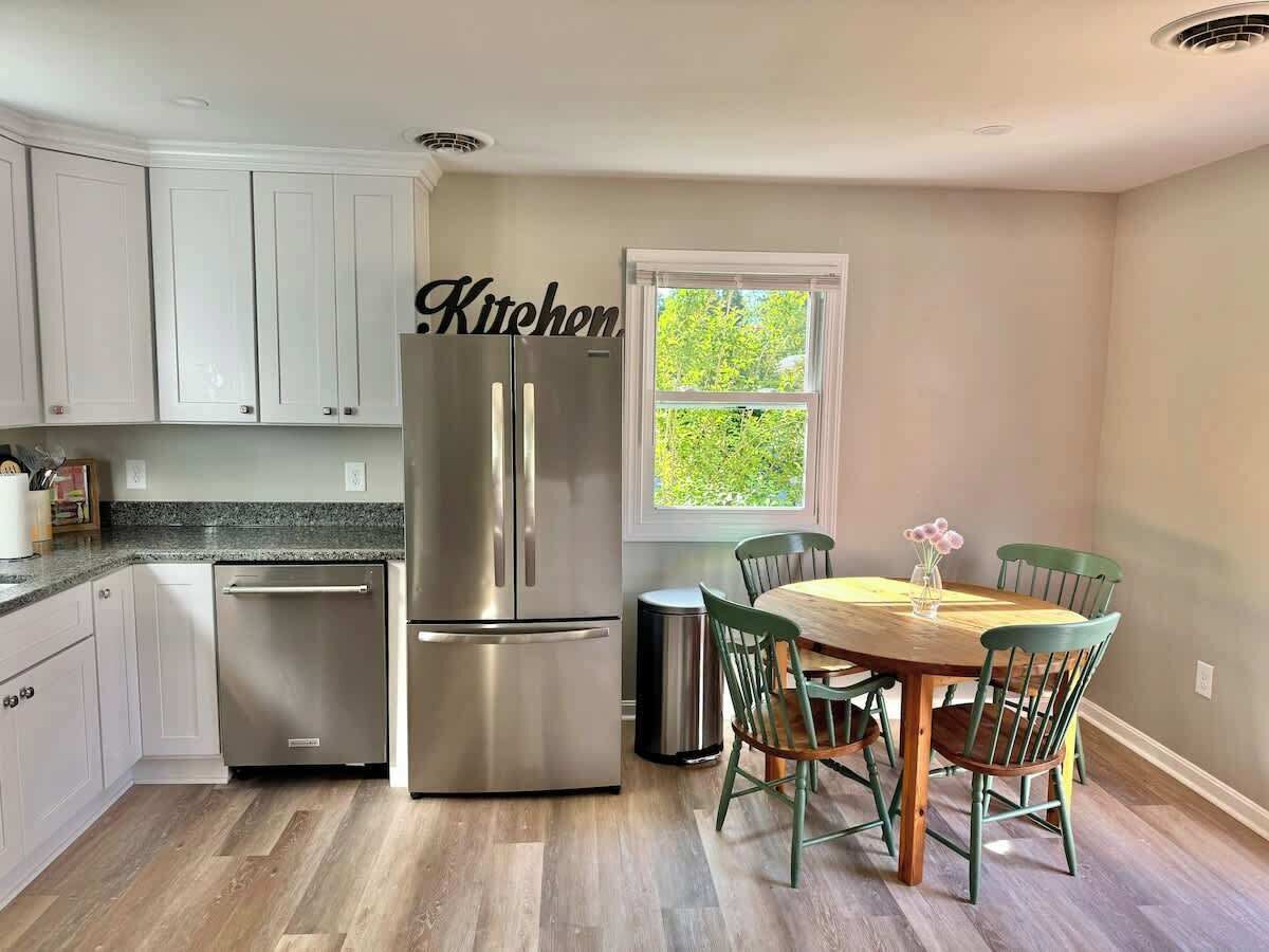 A kitchen with white cabinets, a stainless steel refrigerator, a dining table with chairs, and a window with a view of greenery.