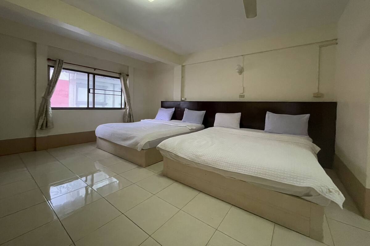 A hotel room with two beds, a ceiling fan, a window with curtains, and a tiled floor.