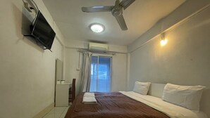 Een hotelkamer met een bed, een plafondventilator, een aan de muur bevestigde televisie en een raam met gordijnen.