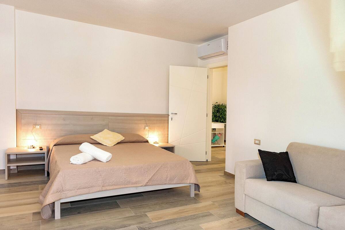 A modern hotel room with a bed, bedside tables, a sofa, and an air conditioning unit.