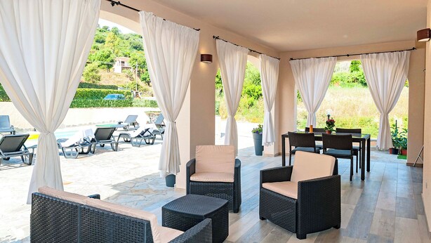 A covered outdoor seating area with wicker furniture and white curtains, overlooking a pool and greenery. - MOLAROTTO CAMERE CON ANGOLO COTTURA (San Teodoro)