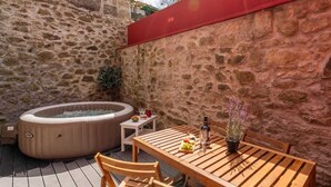 A stone-walled outdoor patio with a wooden table and chairs, a hot tub, and a small white table with a plant and a bottle. - Comfy Flat | Jacuzzi & Terrace by HostWise (Porto)