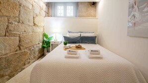 A bedroom with a stone wall, a bed with a white quilt, two pillows, a nightstand with a lamp, a small plant, and a painting on the wall. - Comfy Flat | Jacuzzi & Terrace by HostWise (Porto)