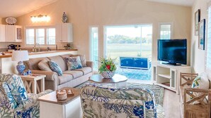A spacious living room with a sofa, coffee table, and TV. A kitchen is visible in the background with cabinets and a window. - Teal Tidez Ocean View Luxury Home w Crows Nest (Murrells Inlet)