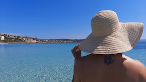 A person wearing a large white hat, facing a clear blue sea with rocky terrain and buildings in the background. - Bosa Marina Fronte Mare (Bosa)