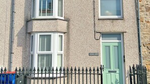 A two-story house with a stone facade, a bay window, a green door, and a black metal fence. - Charming 3 Bed Townhouse Near Llanddwyn Beach (Llanfairpwllgwyngyll)