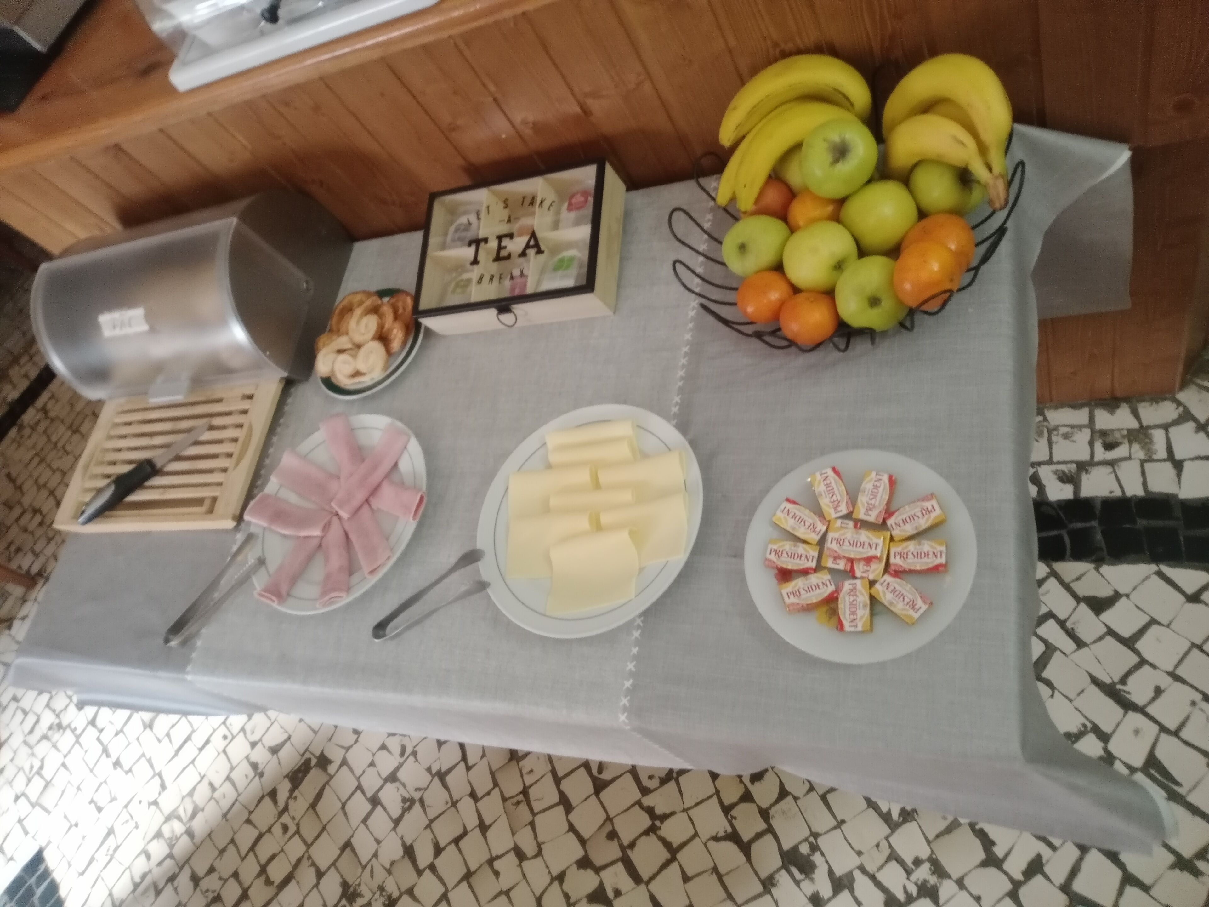 A breakfast table with various food items including bananas, apples, oranges, cheese, ham, and pastries.