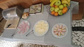 A breakfast table with various food items including bananas, apples, oranges, cheese, ham, and pastries.