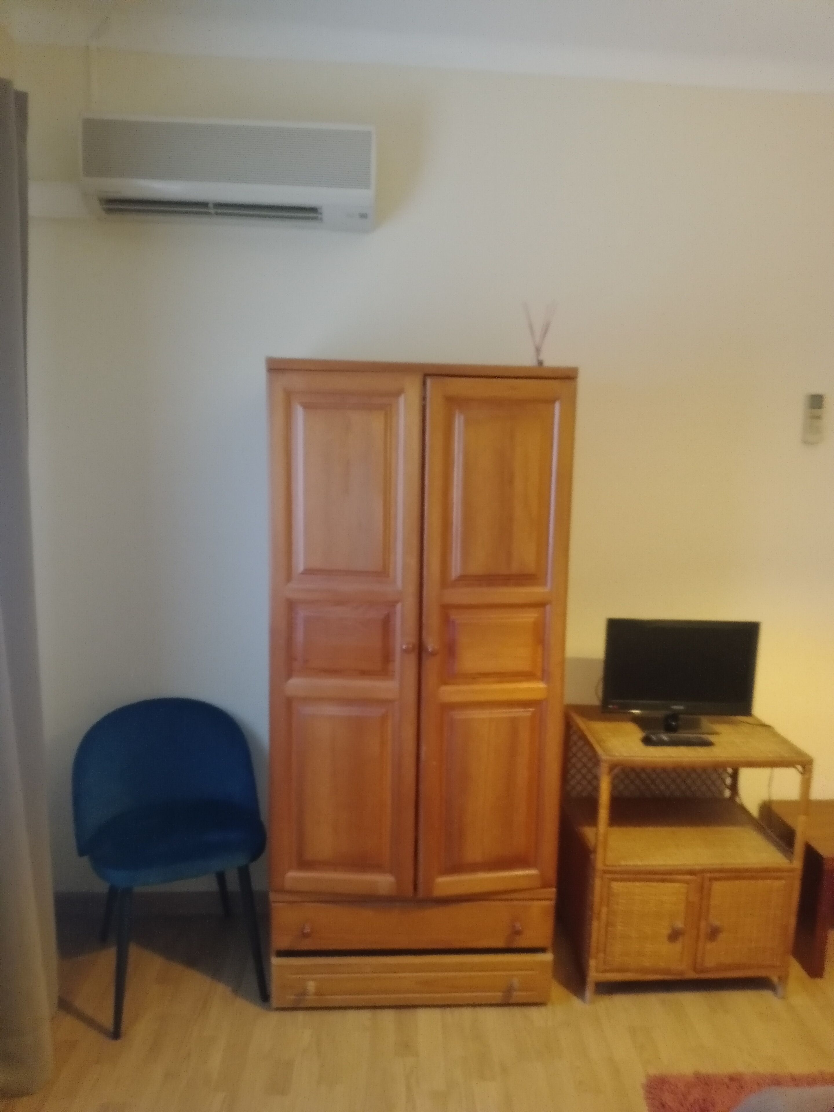 A room with a wooden wardrobe, a blue chair, a television on a small wooden table, and an air conditioning unit on the wall.