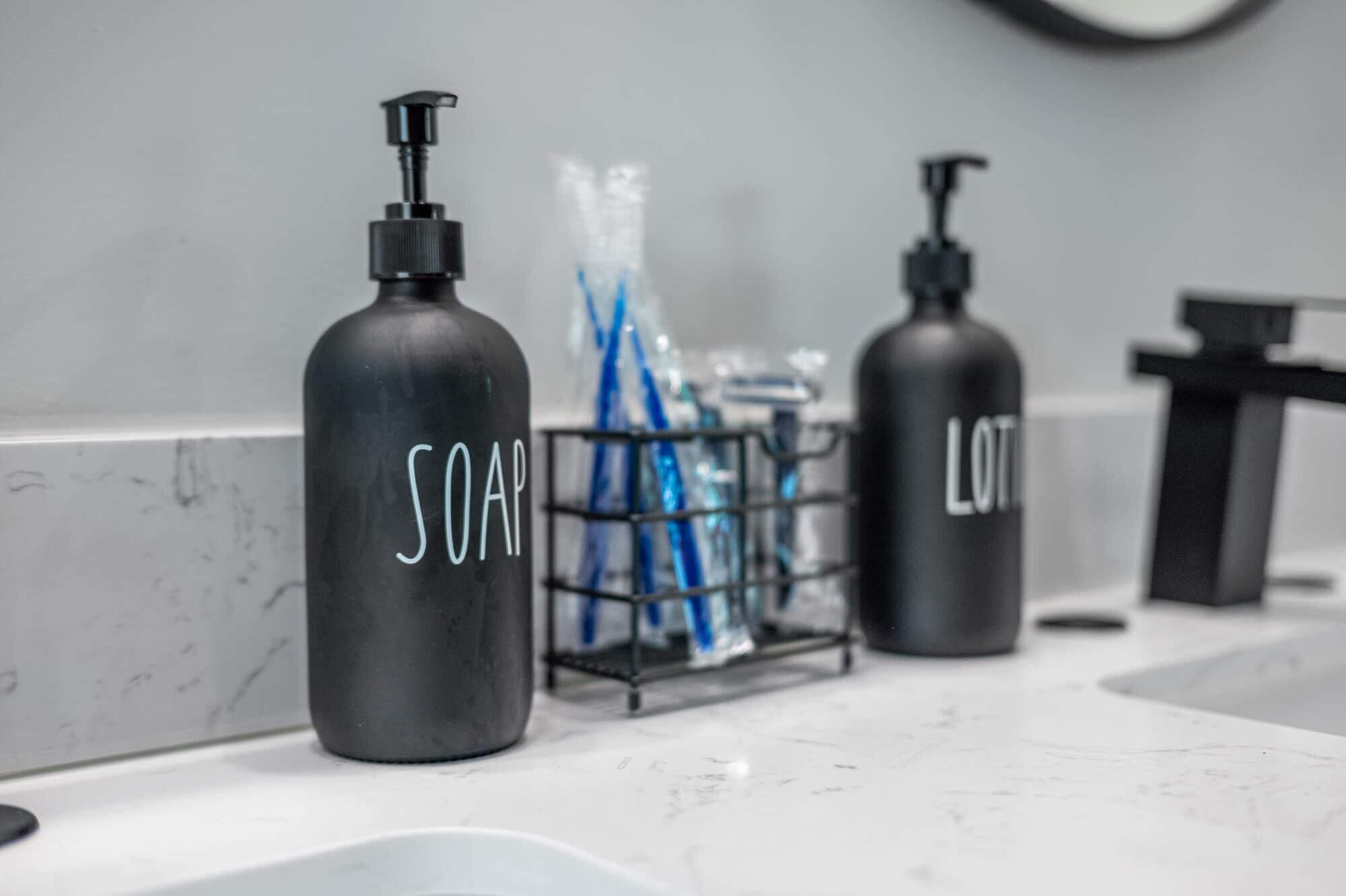 Un baño con encimera de mármol, dos dispensadores de jabón etiquetados 'SOAP' y 'LOT', y un porta cepillos de dientes con cepillos de dientes azules.