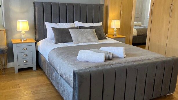 Una cama bien hecha con cabecera gris, dos mesitas de noche con lámparas, y un armario de madera en una habitación de hotel.