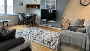 Una sala moderna con televisor de pantalla plana, una zona de comedor con sillas y mesa, un tapete estampado y un rincón acogedor con sofá y sillones.
