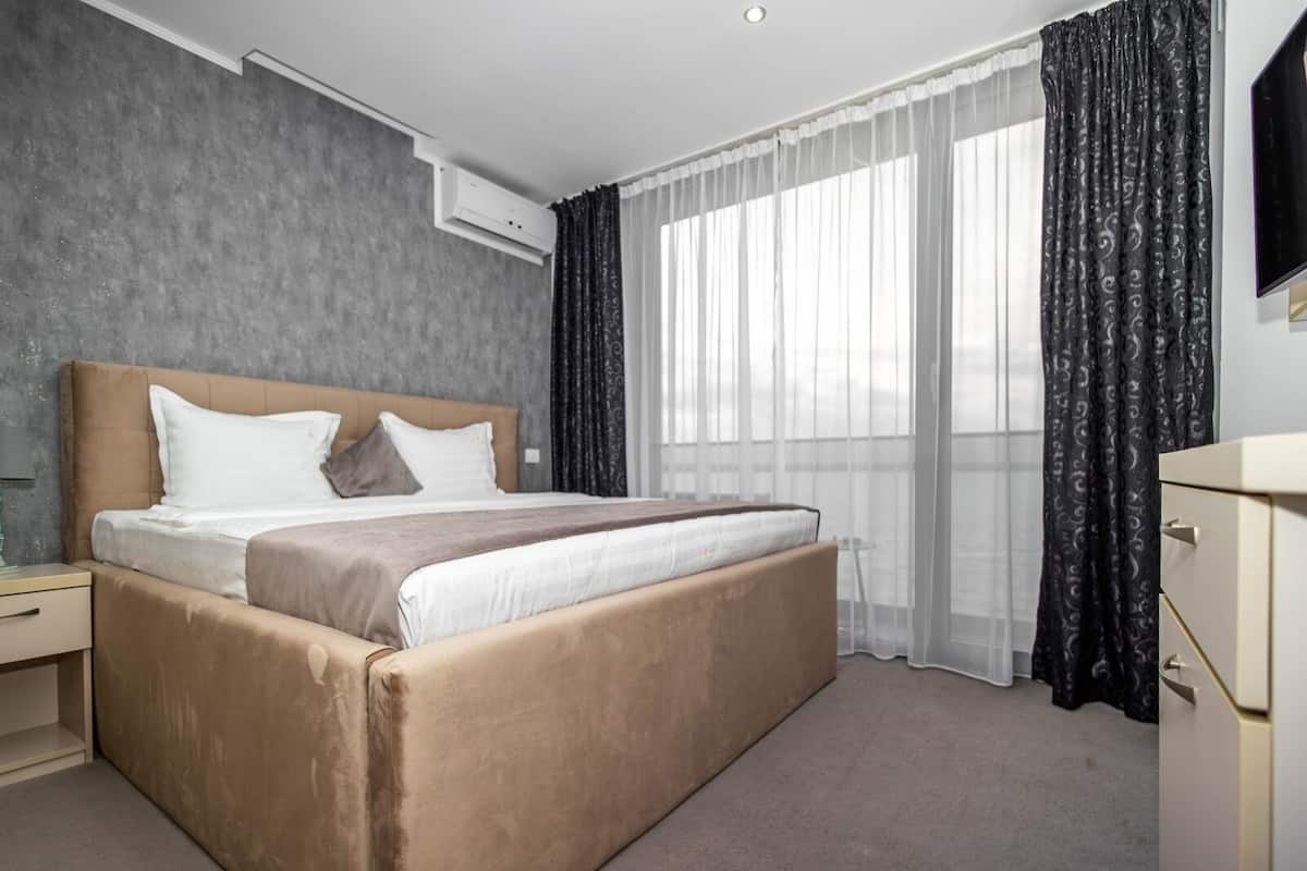 A modern hotel room with a large bed, a bedside table, a TV, and a window with curtains.