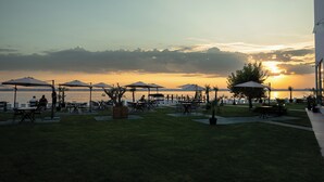 An outdoor seating area with tables and umbrellas, overlooking a body of water and mountains during sunset. - OBA LUXURY BOUTIQUE (Constanta)