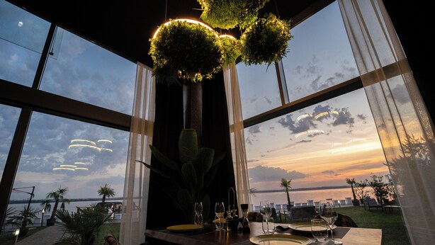 A dining table set for two with a view of the sunset over the water, large windows, and a hanging plant decoration. - OBA LUXURY BOUTIQUE (Constanta)