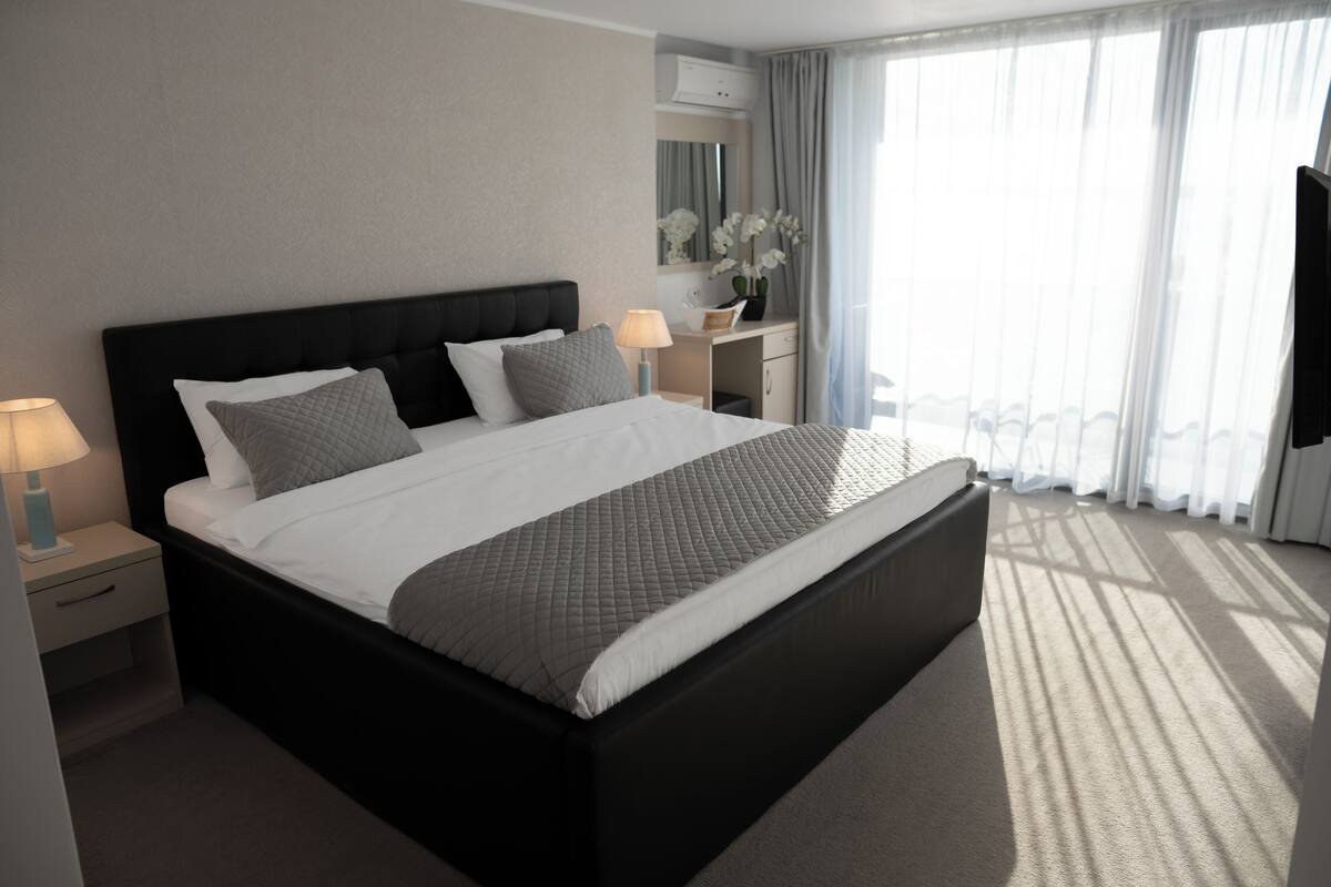 A modern hotel room with a large bed, bedside tables with lamps, a dresser, and a view of the outdoors through sheer curtains.