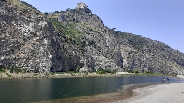 Ein felsiger Hügel mit einem kleinen Gebäude auf der Spitze und ruhigem Wasser im Vordergrund.