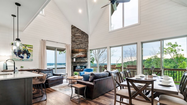 A spacious living area with a stone fireplace, a dining table, and a large window overlooking a deck. - Douglas Lake Life and Pigeon Forge Adventures (Sevierville)