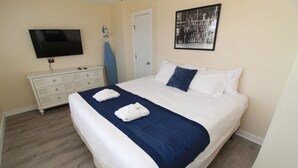 A neatly made bed with a blue blanket and two white towels, a television mounted on the wall, a white dresser, and a framed photo on the wall. - Starfish Suite at Fin & Feather 2BR Condo with Sound Views + Close to Beach (Nags Head)