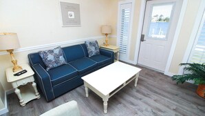 A room with a blue sofa, a white coffee table, a lamp, a framed picture on the wall, a door with shutters, and a potted plant. - Starfish Suite at Fin & Feather 2BR Condo with Sound Views + Close to Beach (Nags Head)