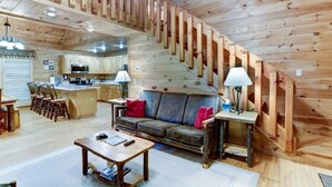 Un interior de cabaña espacioso con escalera de madera, área de cocina, comedor y sala de estar con sofá y mesa de centro.