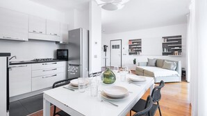 Una cocina moderna con armarios blancos, una mesa de comedor para cuatro personas y una sala de estar con sofá y estanterías.