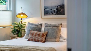 A bedroom with a bed, a bedside lamp, a framed picture of a beach, and a potted plant.