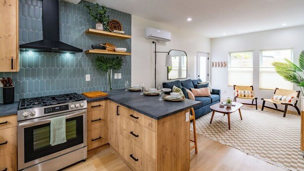 A modern kitchen with a central island, built-in oven, and a black range hood. There is a dining area with a sofa and chairs, and a large window providing natural light.