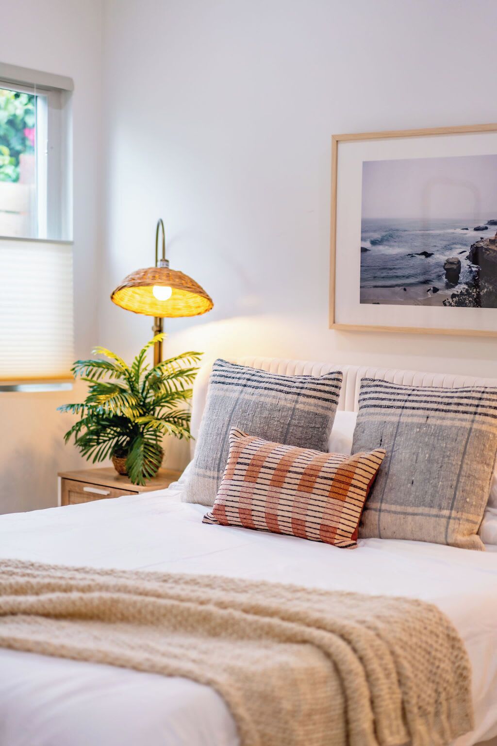 A neatly made bed with a white comforter and multiple pillows, a bedside lamp, a framed picture on the wall, and a potted plant on the nightstand.
