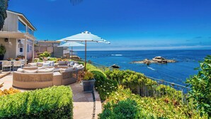 A coastal outdoor seating area with a view of the ocean, featuring a wicker sofa, striped umbrellas, and a pathway leading to a building. - Dolphin's Paradise Su (San Diego)