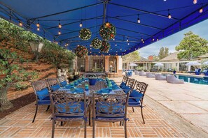 Une terrasse extérieure avec un mur en briques, une piscine et un espace repas couvert, agrémenté de chaises et d’une table bleues.