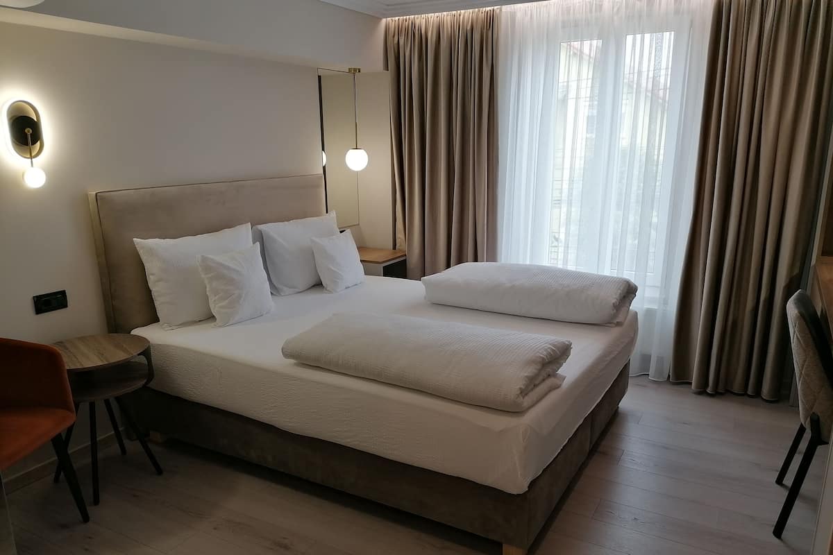 Ein modernes Hotelzimmer mit einem großen Bett, einem Nachttisch, einem Sessel und einem großen Fenster mit Vorhängen.