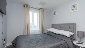 Une chambre à coucher avec un lit gris, une tête de lit grise, une table de chevet avec une lampe et une fenêtre avec des rideaux.