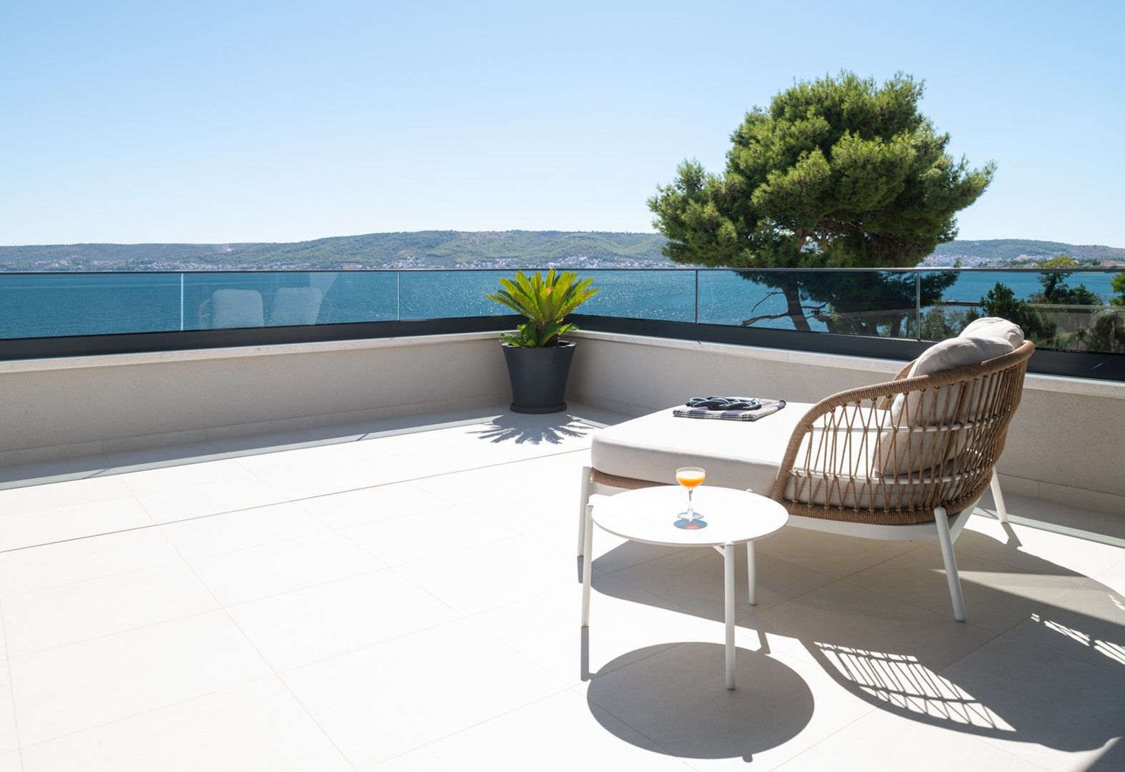 Ein Balkon mit Meerblick, ein Blumentopf, ein Korbstuhl, ein kleiner Tisch mit einem Getränk und ein weißes Sofa.