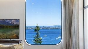 A room with a large window offering a view of a lake with boats, a tree, and a distant cityscape.