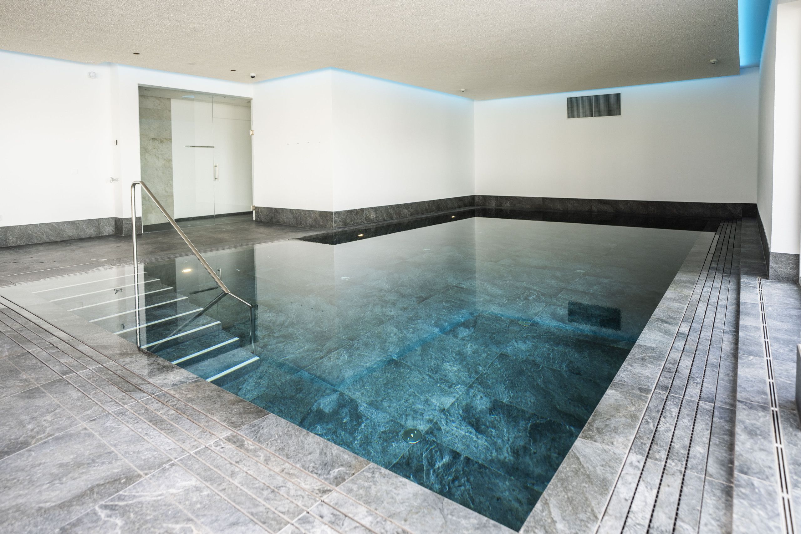 Ein Hallenbad mit glasklarer Wasseroberfläche, einer Glasbrüstung und einem gefliesten Boden.