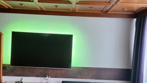 A room with a wooden ceiling, a large flat-screen TV mounted on the wall, a wooden desk with a chair, a dark sofa, and a red carpet. - Eibacher Hof (Nuremberg)