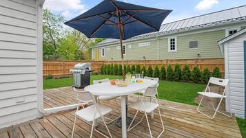 Une terrasse en bois avec une table et des chaises blanches, un grand parasol et un barbecue.