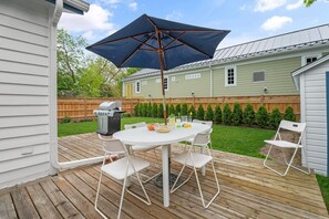 Una terraza de madera con una mesa y sillas blancas, un paraguas grande y una parrilla.