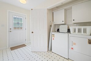 A compact kitchen with a white door, a window with blinds, a washing machine, and a dryer. - NEW! The Cedar House - Steps from Queen Street (Niagara-on-the-Lake)