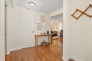 A hallway with wooden flooring, a white door, a small table with items on it, and a wicker basket. - NEW! The Cedar House - Steps from Queen Street (Niagara-on-the-Lake)